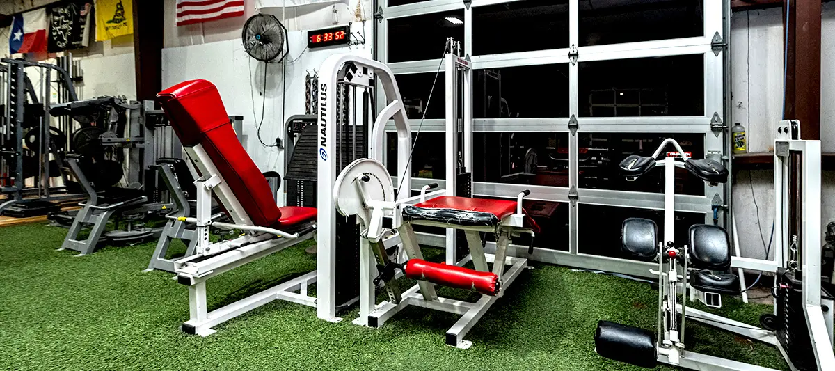 Rows of plate-loaded and selectorised machines on turf, with a roll-up door letting in daylight behind them.