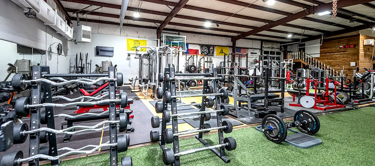 A wide view of the gym floor: racked barbells and dumbbells in the foreground, power racks, benches and platforms beyond, under a high metal roof.